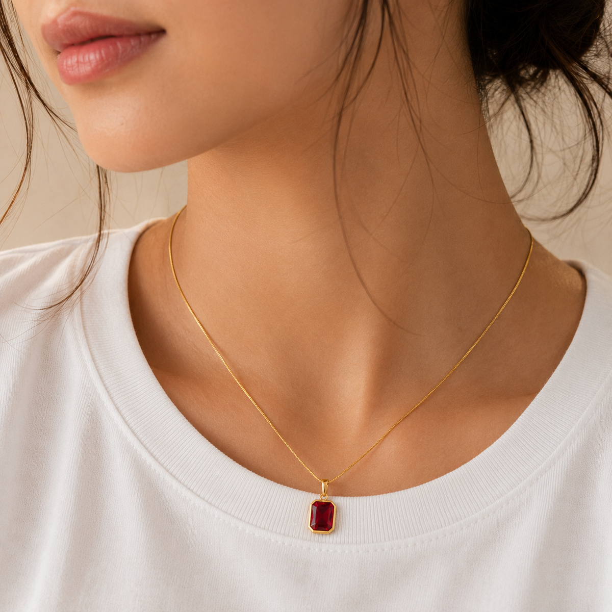 A close-up of a model wearing a delicate gold chain necklace with a small rectangular red gemstone pendant, paired with a white crew-neck t-shirt.