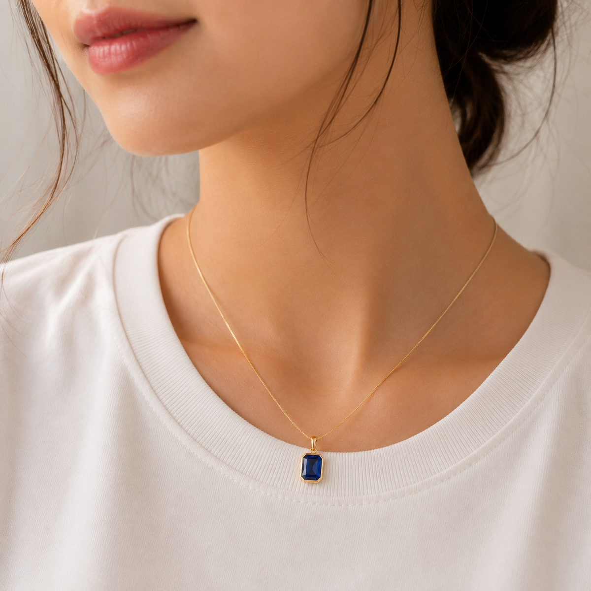 A close-up of a model wearing a delicate gold chain necklace with a rectangular dark blue gemstone pendant, styled with a white crew-neck t-shirt.