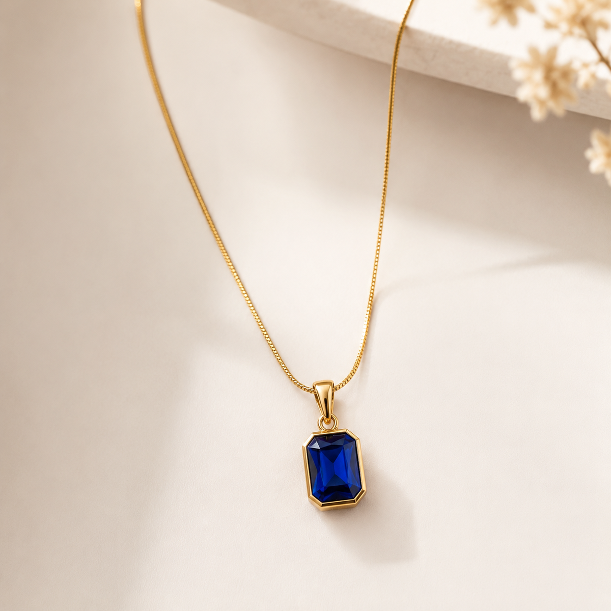 A gold chain necklace with a rectangular blue gemstone pendant, displayed on a light beige surface with soft shadows and dried flowers in the corner.