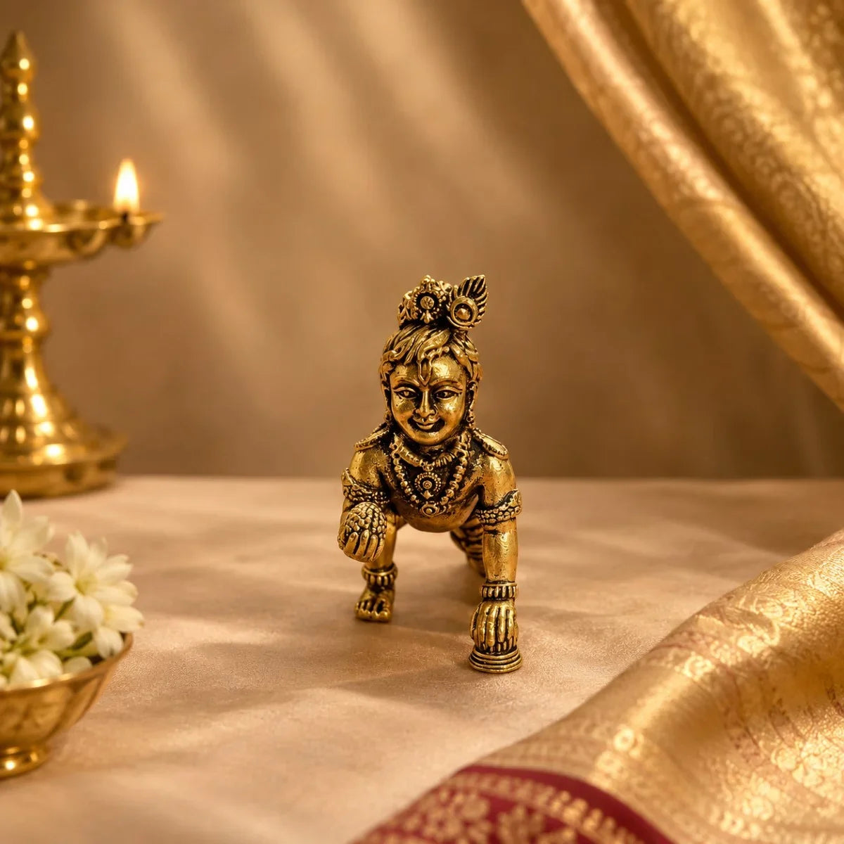 Brass Laddu Krishna crawling idol with diya and jasmine flowers on gold drape. Zishta Janmashtami Gifts.