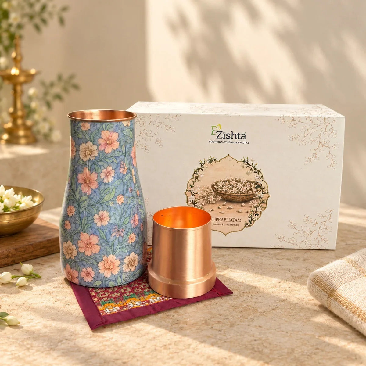 Blue floral enamel copper water carafe with copper tumbler on a Madur grass coaster, with the Zishta Suprabhatam gift box behind. Zishta Diwali Corporate Gifting.