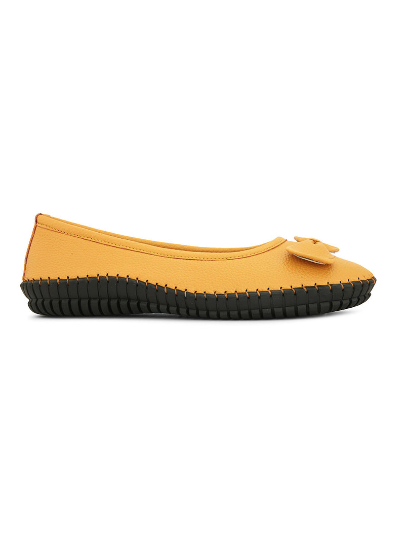 Mustard yellow leather flat shoe with a decorative bow, black textured sole, and visible stitching for a casual, stylish look.