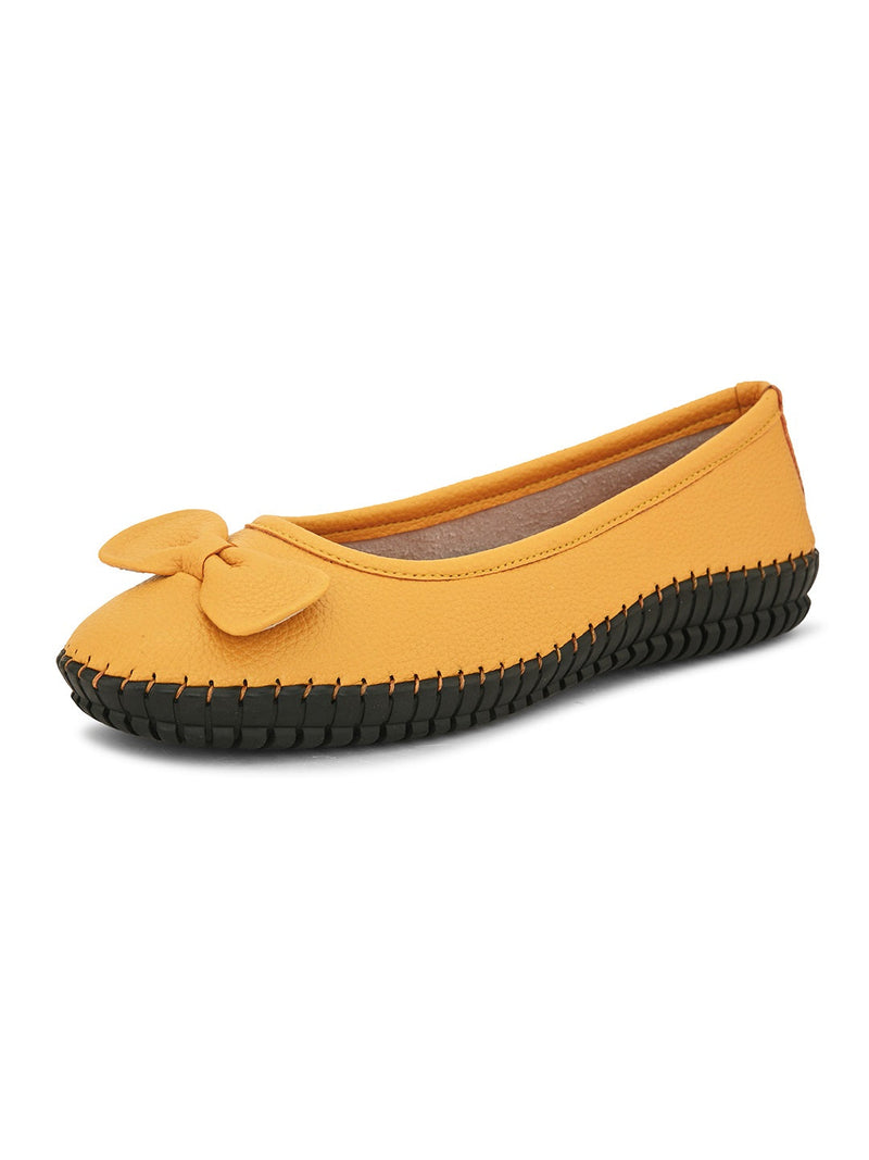 Mustard yellow leather flat shoe with a decorative bow, black textured sole, and visible stitching for a casual, stylish look.